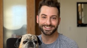 A smiling man with short dark hair and a dark beard is wearing a grey sweater and holding a black and tan pug.