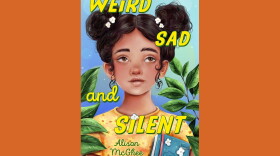 Cover of "Weird Sad and Silent" by Alison McGhee; colorful illustration of a girl in a yellow-orange floral shirt, holding a book and standing between two green plants