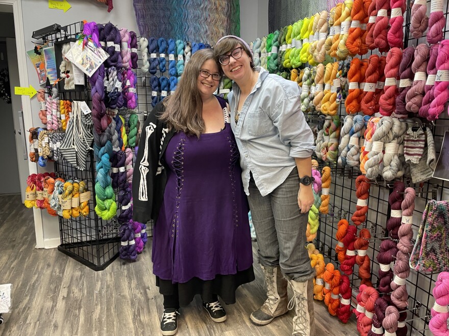 Two women lean into each other, smiling, in front of a wall with lots of colorful yarn hanging from racks
