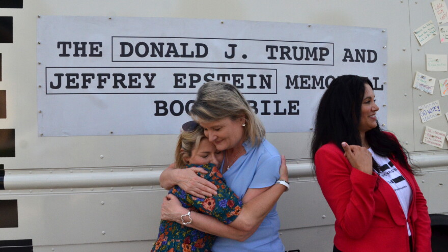 Two women hug in front of a truck that reads "The Donald J. Trump and Jeffrey Epstein Memorial Bookmobile."