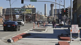 Exterior photo of a busy street at left. At right two people are walking away from the camera on a freshly poured concrete space.