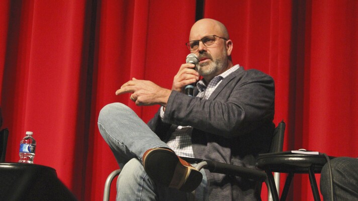 A man wearing glasses, a blazer and jeans sits onstage in front of a red curtain, holding a microphone and gesturing as he speaks. A small table with a water bottle sits beside him.