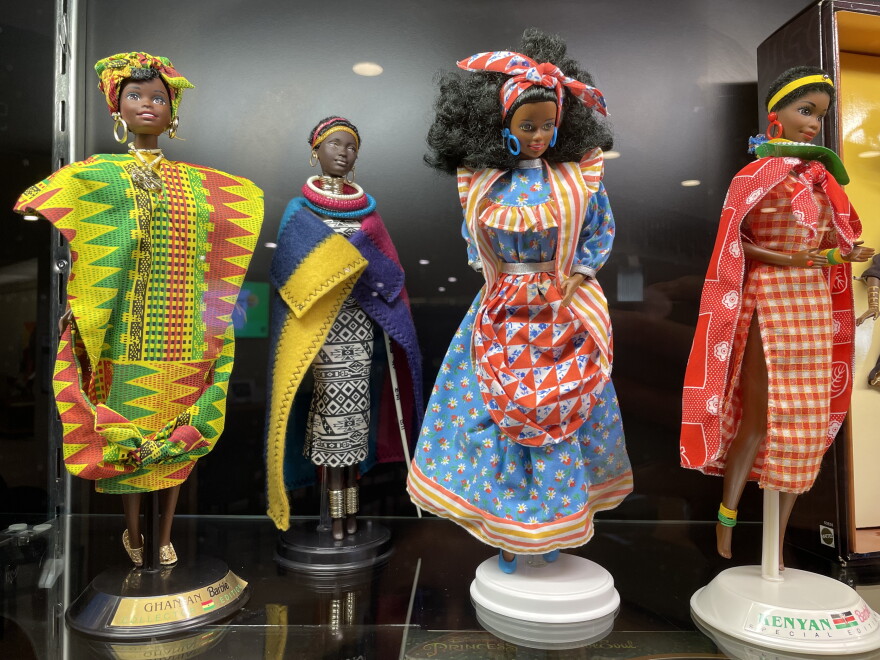 Four special edition Black Barbie dolls are pictured standing in a case. They represent African and Caribbean countries in their dress.