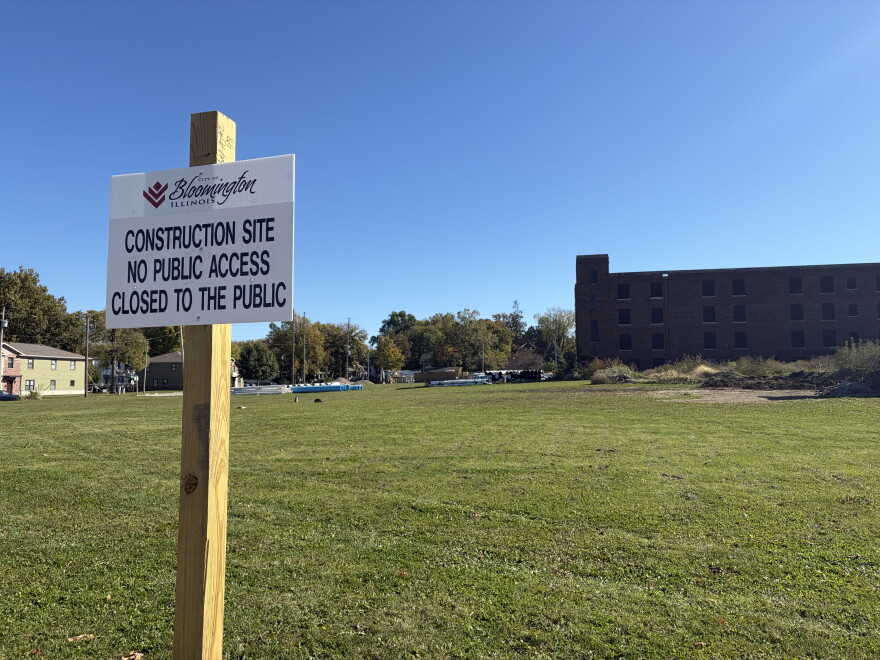 in the foreground, a white sign on a wooden post. in the background, an open field and brown brick building