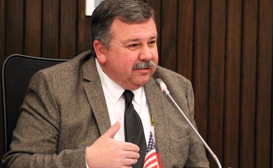 A man in a gray suit jacket and black tie speaks into a microphone at a meeting. He is sitting at a table with a small American flag in front of him and a dark wood-paneled background behind.