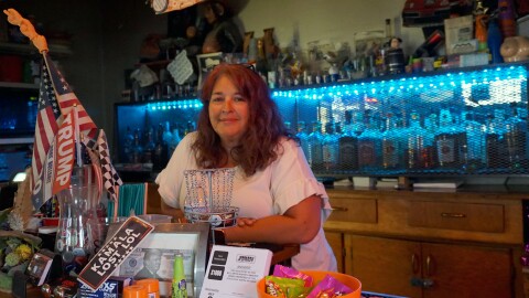 A woman with red/brown hair and a white shirt leans against a bar, with a shelf of liquor in the background and pro-Trump signs in the foreground.