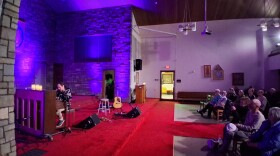A person playing piano on a low, red-carpeted stage in front of a seated crowd