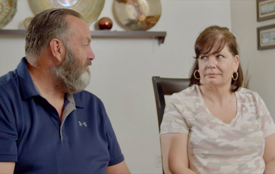 A man in a blue shirt gazes and a woman in an off-white shirt look at one another.