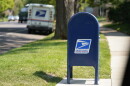 A blue USPS mailbox surrounded by green grass on a residential street. A USPS mail truck is visible in the distance.