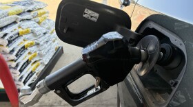 A black gas pump nozzle is inserted into a gray SUV with a pallet of bags of ice melting compound visible in the background. 