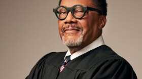 Judge Gregory Mathis stands in a black judge's robe with his arms crossed. He's wearing black framed glasses.