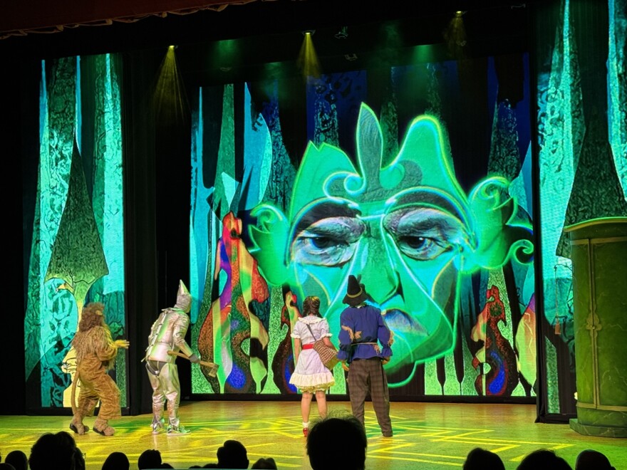 People dressed as Wizard of Oz characters stand on a stage looking at an on-screen representation of the Wizard.