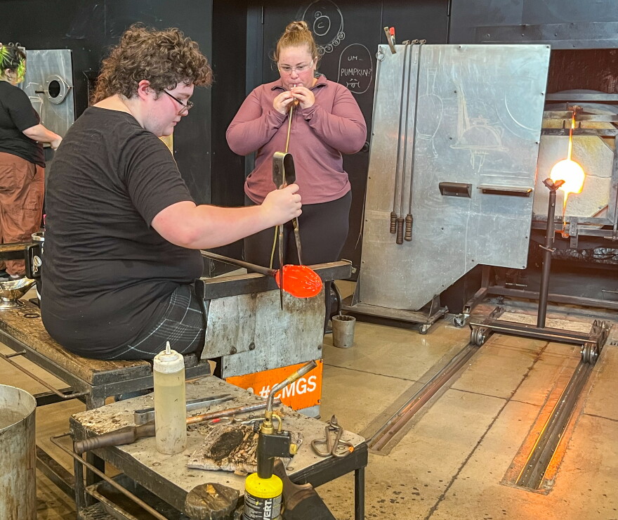 Perry Glass Studio assistant Jal Pierce, seated, holds a tube of molten glass that Jenna Schatz is inflating with a blowpipe. Pierce is helping Schatz make a glass pumpkin.