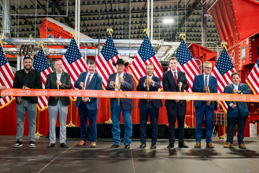 Men in a factory stand in front of a ribbon holding scissors about to cut it.
