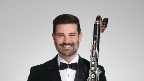 Man with close-cropped dark hair, a beard, and a mustache, dressed in a black tuxedo, holds a bass clarinet in his left arm. 