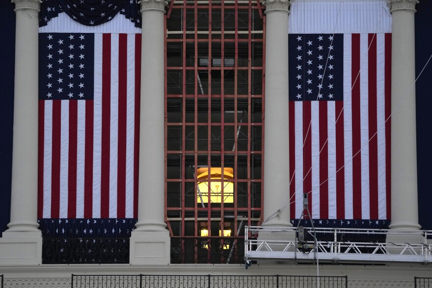 Flags hang in place on the West Front of the U.S. Capitol, with a metal platform hanging in front of one of the flags. A large window bisects the two flags, showing a glimpse inside the Capitol Building. 