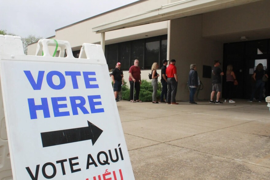 A sign that says "Vote Here" stands outside a polling place where people stand in line. 