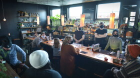 Nine people, all wearing surgical-type masks, gather around a restaurant bar. Some stand some sit. Lots of bottles of liquor and stemware are seen in the background.