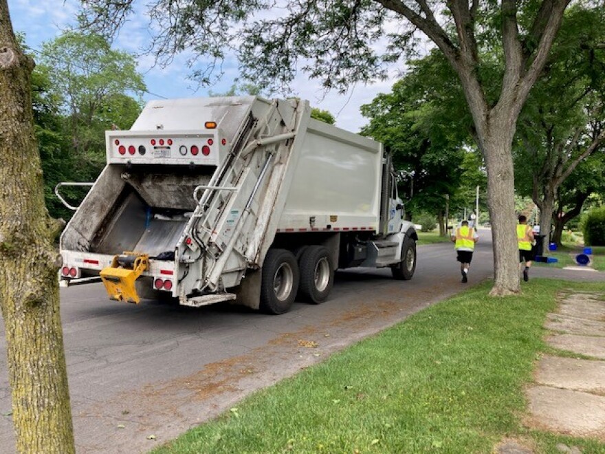 A trash truck rolls down a street lined with trees as two workers run to the next house.