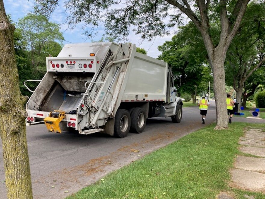  A trash truck rolls down a street lined with trees as two workers run to the next house.