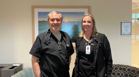 A man and woman pose for a picture in the lobby of a clinic. They are both wearing black scrubs.