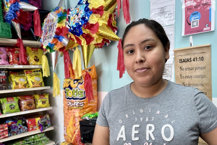 A young Hispanic woman stands in her party shop with a bible verse in the background