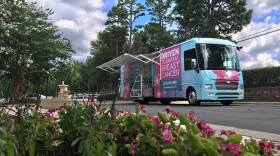 Pink flowers are in the foreground. Behind them is a street and parking lot with a bus parked in it. The bus is light blue and decorated with pink flowers. A large pink square on it reads "Driven to defeat breast cancer." A collapsible awning is opened on the bus's side where patients will step into the vehicle for a mammogram.