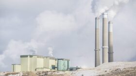 The stacks of a large power plant tower over a hilly snow-covered ground.