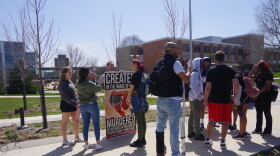 A man stands behind a giant anti-abortion sign as he talks to students on campus.  Most of the students are heckling him, politely.  