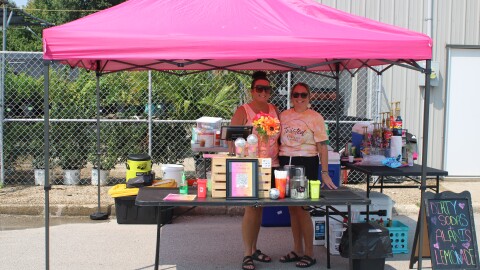 Two women stand in a pop-up tent where they are serving dirty sodas