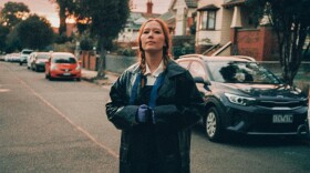  A woman wearing a long trenchcoat stands in the middle of a residential street with cars parked along the curb and a row of houses behind her.
