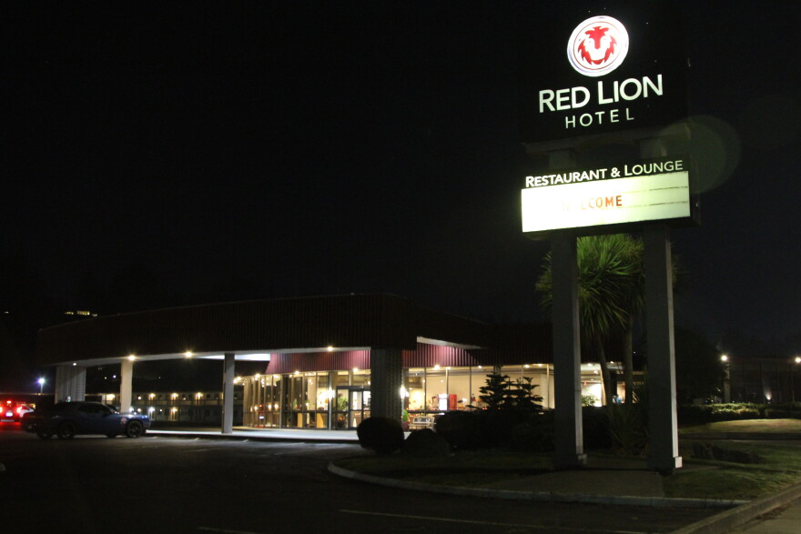 The lights through a hotel lobby window illuminate a dark parking lot at nighttime and a large sign with the words, "Red Lion Hotel." 