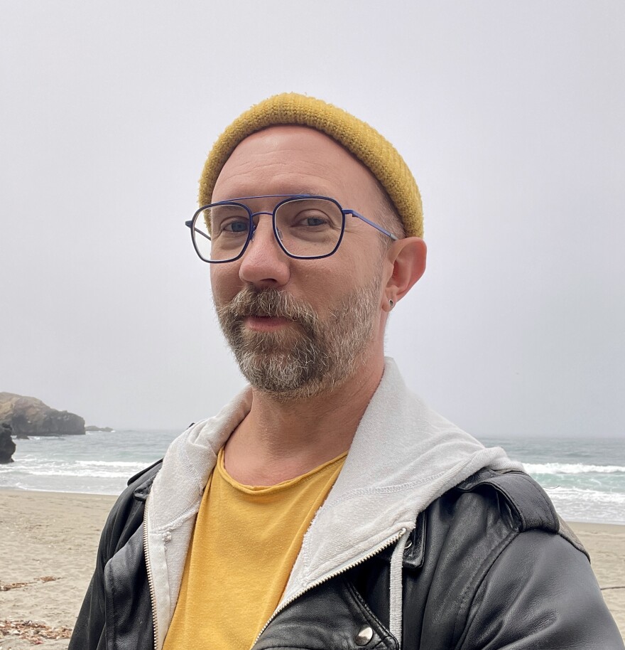Nathan Hall, a man wearing a yellow Tshirt and knitted stocking cap, a white hoodie and a black leather jacket, stands on a beach with the ocean and a gray sky behind him.