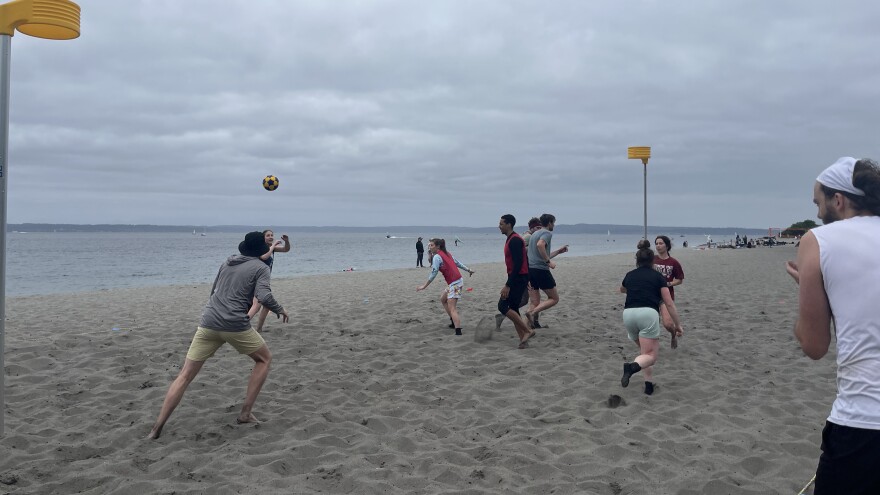 People throw a ball up and run around on a beach on a cloudy day.