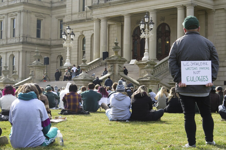 People sit on the lawn outside the state capitol. One holds a sign that says, 'we should be writing papers, not eulogies.'