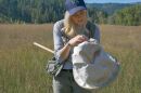 A blonde haired woman stands in a grassy meadow with an insect net. 