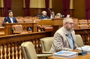 A man with a beard and glasses sits at a table in a courtroom, with other people seated in the background.