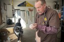 A man with a stethoscope pulls medication out of a bottle with a needle. Barn supplies surround him and a black and white cat stands next to him.