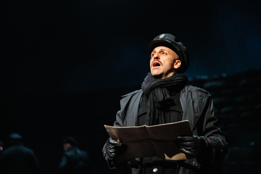A man in a black overcoat and black military hat holds a notebook. He is singing.