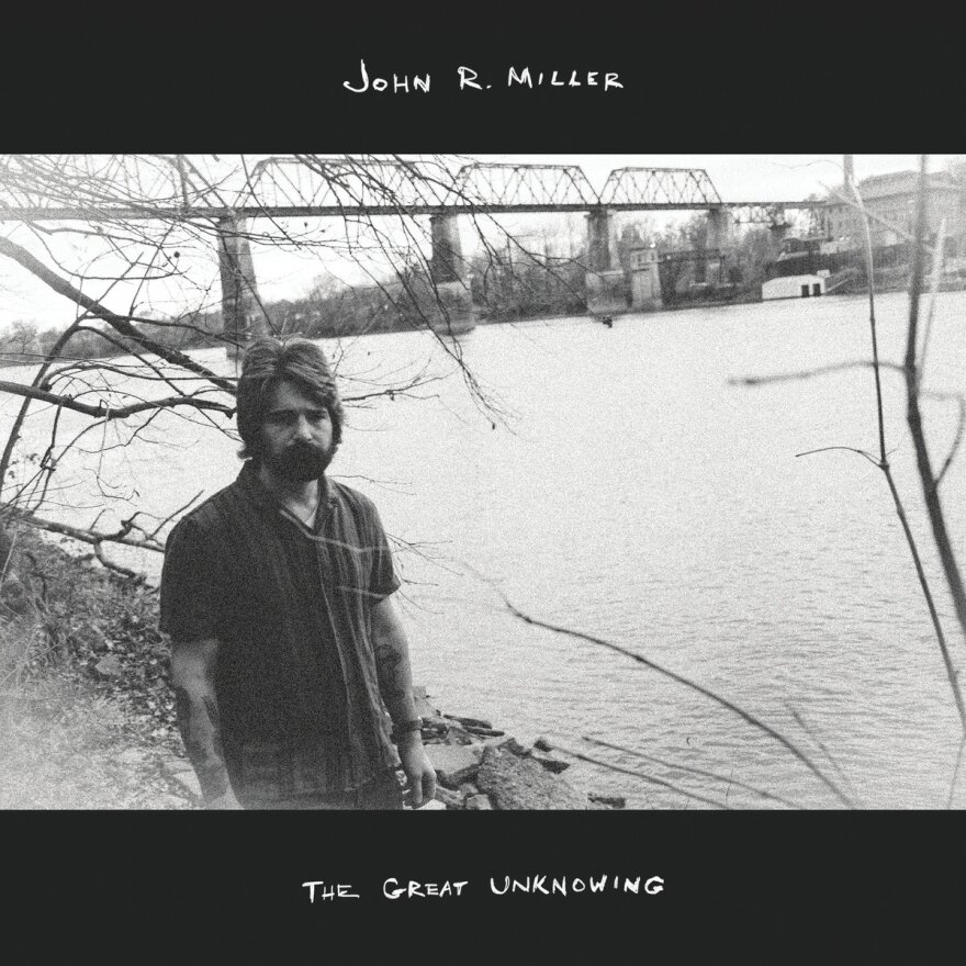 A bearded man stands next to a body of water in a black and white image.