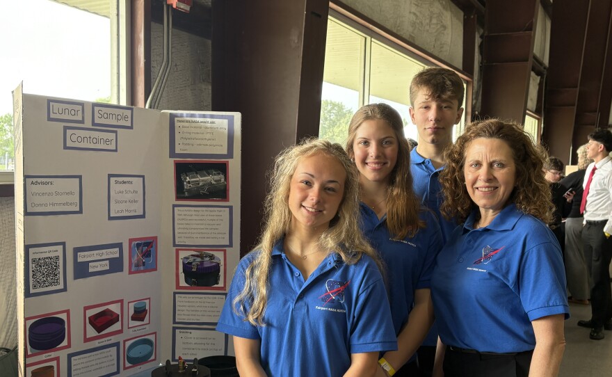 Four people wearing bright blue polo shirts stand in front of a posterboard presentation.