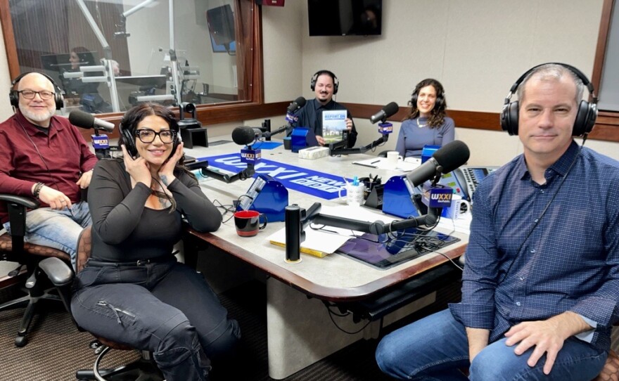 Five smiling people wearing headphones sit at a table in a radio talk studio. A man front left is bald and has a white beard; he is wearing glasses, a red sweater, and jeans. A woman front center has long brown hair and is wearing glasses, a long-sleeved grey shirt, black jeans, and black shoes; a man at right has short dark hair and is wearing a blue checked button-down shirt, jeans, and brown shoes; a man back left has short black hair and a black mustache and is wearing a dark shirt; a woman back center has curly brown hair and is wearing a necklace and a long-sleeved blue shirt.
