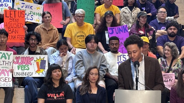 A man speaks at a podium as a crowd behind him holds signs protesting "junk fees."