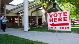 A "vote here" sign staked in the grass in front of some red picnic tables.
