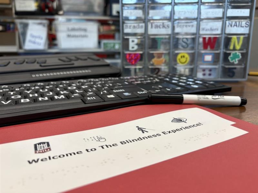 A red folder sits on a desk, surrounded by a marker, a large-print computer keyboard, and a storage bin that visually-impaired people can use for hardware. One of the rows is labeled "nails" and features a textured letter "n" and an actual nail glued onto the front so they could distinguish what's placed inside. The red folder reads "Welcome to the Blindness Experience!" and says the same underneath in braille. 