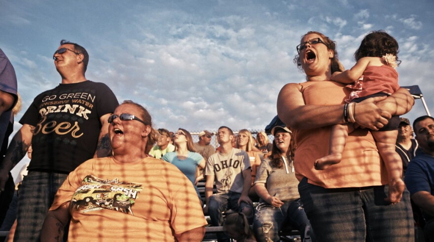 People cheer on outdoor bleachers on a sunny evening. 
