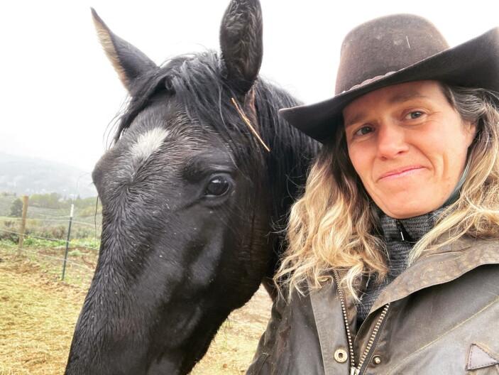 Ashley Ahearn, wearing a cowboy hat and jacket, takes a selfie with black mustang with a white spot on his head.
