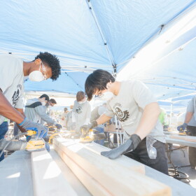 Volunteers wearing masks and safety glasses use power sanders on lumber beneath outdoor canopies.