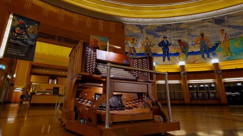 A complicated-looking wooden organ, with a multitude of pedals, knobs and keys, sits in the middle of a grand room. There is a mural in the background.