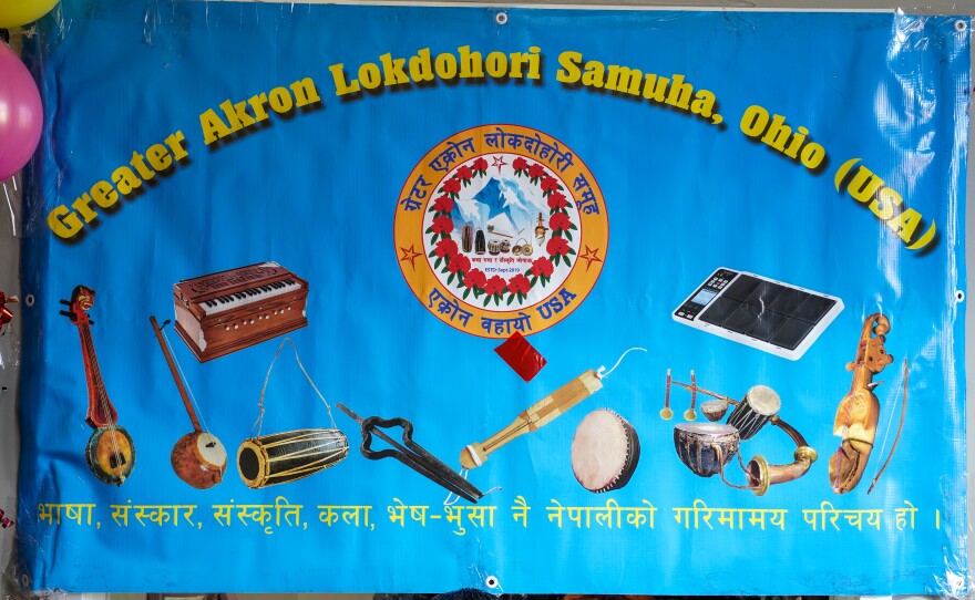 Large blue sign that has pictures of Nepalese musical instruments and reads "Greater Akron Lok Dohori Samuha" in English with Nepalese script at the bottom.